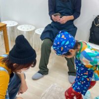 カラフルなエプロンの男の子が透明シート越しのうどん生地を足で踏み、お母さんが近くで見守っている場面 手打ちうどん作り体験教室 2025年11月 01.jpg – イリコスキー製麺所