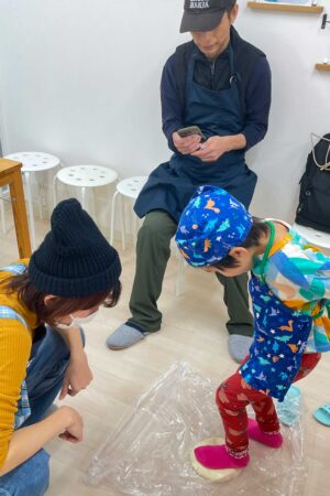 カラフルなエプロンの男の子が透明シート越しのうどん生地を足で踏み、お母さんが近くで見守っている場面 手打ちうどん作り体験教室 2025年11月 01.jpg – イリコスキー製麺所