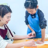 青いエプロンの女の子が麺切包丁でうどん生地を切り、お母さんが手元を支えている様子 手打ちうどん作り体験教室 2025年11月 02.jpg – イリコスキー製麺所