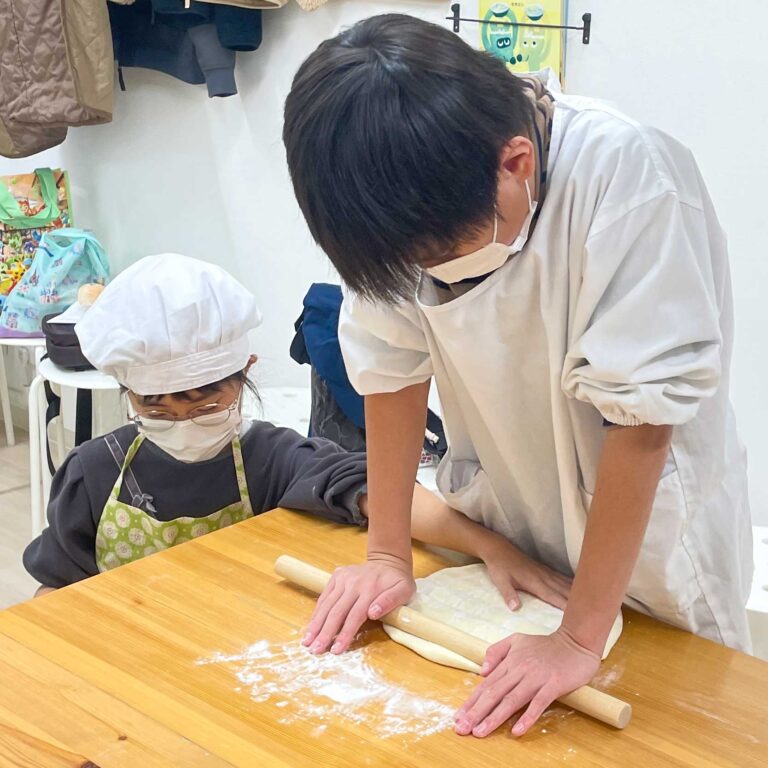 妹が見守る中、お兄ちゃんが麺棒でうどん生地を大きく伸ばしている場面 手打ちうどん作り体験教室 2025年8月 2.jpg – イリコスキー製麺所