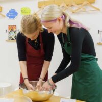 赤いエプロンと緑のエプロンの女性2人が、ボウルの中で生地作り(粉と水で生地を作る工程)をしている 手打ちうどん作り体験教室 2025年12月 A6407348-1-scaled.jpg – イリコスキー製麺所