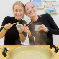 女性2人が湯気の立つ鍋の前で、箸と蕎麦猪口を持ってうどんを味わっている 手打ちうどん作り体験教室 2025年12月 A6407372-1-scaled.jpg – イリコスキー製麺所