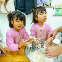 ピンクのエプロンの子ども2人が大きなボウルで粉と水を混ぜて生地を作り、横から大人の手がやさしく手伝っている 手打ちうどん作り体験教室 2025年12月 A6407452-1-scaled.jpg – イリコスキー製麺所