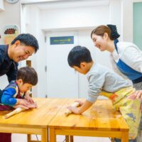 木のテーブルで、幼い子どもが麺棒を転がし、大人が近くで手を添えてサポートしているうどん作り体験の様子 手打ちうどん作り体験教室 2025年12月 A6407476-1-scaled.jpg – イリコスキー製麺所