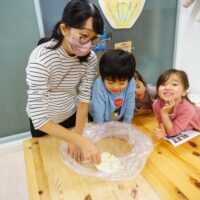 テーブルで生地を扱う大人のそばで、子どもたちが笑顔で見守りながら作業を楽しんでいる教室の風景 手打ちうどん作り体験教室 2025年12月 A6407486-1-scaled.jpg – イリコスキー製麺所