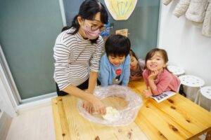 テーブルで生地を扱う大人のそばで、子どもたちが笑顔で見守りながら作業を楽しんでいる教室の風景 手打ちうどん作り体験教室 2025年12月 A6407486-1-scaled.jpg – イリコスキー製麺所