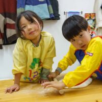 黄色のスモックを着た二人の子供が麺棒を使って生地作りをしている場面 / Two children in yellow smocks using a rolling pin to work on the dough. 手打ちうどん作り体験教室 2025年12月 A6407641-1-scaled.jpg – イリコスキー製麺所