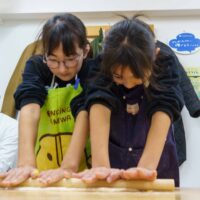 二人の女の子が並んで力いっぱい麺棒でうどん生地を伸ばしている様子 / Two girls side-by-side using all their strength to roll out udon dough with a rolling pin. 手打ちうどん作り体験教室 2025年12月 A6407647-1-scaled.jpg – イリコスキー製麺所
