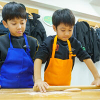 真剣な表情で並んで麺棒を動かし生地作りを行う男の子たち / Boys working side-by-side with serious expressions, using rolling pins to make udon dough. 手打ちうどん作り体験教室 2025年12月 A6407728-1-scaled.jpg – イリコスキー製麺所