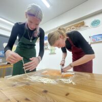 緑のエプロンの女性が切ったうどんを持ち上げ、赤いエプロンの女性が麺を切っている 手打ちうどん作り体験教室 2025年12月 IMG_1074-scaled.jpg – イリコスキー製麺所