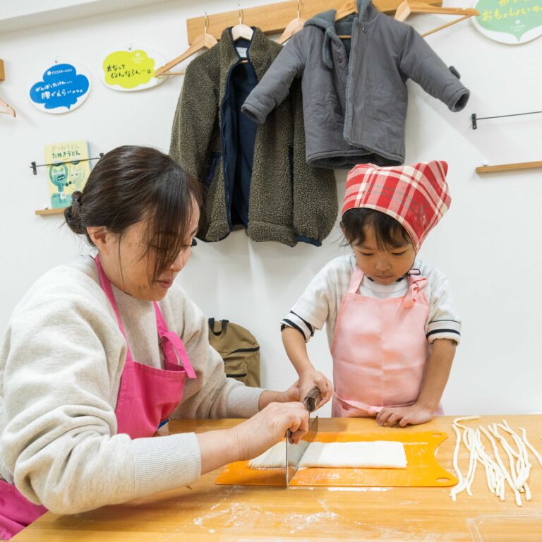 真剣な表情でうどんの生地を包丁で切る女の子と女性 (A girl and her mother cutting udon dough with a knife with serious expressions) 手打ちうどん作り体験教室 2025年12月 4-1.jpg – イリコスキー製麺所
