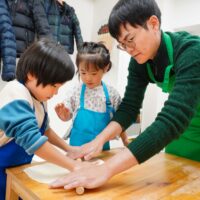 男性が子供たちの手元に添えて一緒に麺棒で生地を薄く伸ばしている様子。 / A man helping children roll out the dough thinly with a rolling pin. 手打ちうどん作り体験教室 2026年1月 A640733-1-scaled.jpg – イリコスキー製麺所