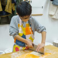 色鮮やかなエプロンを身につけ、真剣な表情でうどんを包丁で切る男の子。A boy in a colorful apron carefully cutting udon noodles with a knife. 手打ちうどん作り体験教室 2026年1月 A6407790-1.jpg – イリコスキー製麺所