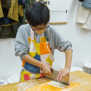 色鮮やかなエプロンを身につけ、真剣な表情でうどんを包丁で切る男の子。A boy in a colorful apron carefully cutting udon noodles with a knife. 手打ちうどん作り体験教室 2026年1月 A6407790-1.jpg – イリコスキー製麺所