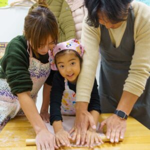ピンクのバンダナを巻いた女の子が両親と一緒に麺棒を転がしている手元。A young girl in a pink bandana rolling out the dough with her parents' help. 手打ちうどん作り体験教室 2026年1月 A6407810-1.jpg – イリコスキー製麺所