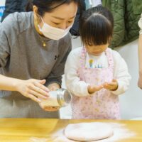 ピンク色の生地に打ち粉を振る手元を、女の子が慎重に見守る場面 / A young girl carefully watching as flour is sprinkled onto pink dough. 手打ちうどん作り体験教室 2026年1月 A6407818-1-scaled.jpg – イリコスキー製麺所