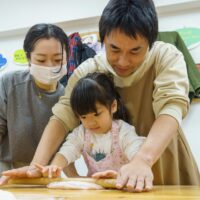 麺棒をしっかりと握り、体重をかけてピンク色の生地を伸ばす女の子 / A girl firmly grasping a rolling pin and putting her weight into stretching the pink dough. 手打ちうどん作り体験教室 2026年1月 A6407825-1-scaled.jpg – イリコスキー製麺所