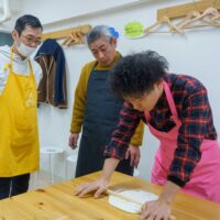 ピンクのエプロン姿で生地を伸ばす様子を真剣に見つめる男性ゲストたち / Male guests watching intently as a man in a pink apron rolls out the dough. 手打ちうどん作り体験教室 2026年1月 A6407912-1-scaled.jpg – イリコスキー製麺所