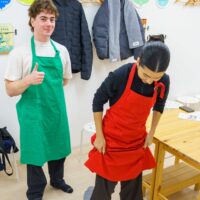 ビニール袋に入れた生地を足で踏む女性と、笑顔で親指を立てる男性。 / A woman stepping on udon dough in a plastic bag while a man gives a thumbs-up with a smile. 手打ちうどん作り体験教室 2026年1月 A6407924-1-scaled.jpg – イリコスキー製麺所