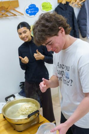 A6407933-1.jpg：茹で上がるうどんを前に、満足げにポーズをとるお二人。 / Two guests posing happily in front of the boiling udon. 手打ちうどん作り体験教室 2026年1月 A6407933-1.jpg – イリコスキー製麺所