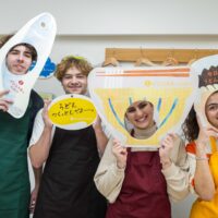 うどんや魚のイラストが描かれた顔はめパネルを持って楽しむ4人の若者 Four young people having fun holding comic foregrounds (kao-hame panels) with illustrations of udon and fish 手打ちうどん作り体験教室 2026年1月 A6408146-1-scaled.jpg – イリコスキー製麺所