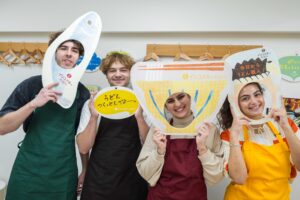 うどんや魚のイラストが描かれた顔はめパネルを持って楽しむ4人の若者 Four young people having fun holding comic foregrounds (kao-hame panels) with illustrations of udon and fish 手打ちうどん作り体験教室 2026年1月 A6408146-1.jpg – イリコスキー製麺所