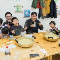 大きな鍋のうどんを囲み、箸やピースサインで笑顔を見せる2組の友人グループ alt: Two groups of friends smiling with chopsticks and peace signs around large pots of udon. 手打ちうどん作り体験教室 2026年1月 A6408189-1-scaled.jpg – イリコスキー製麺所