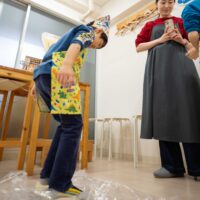 ビニール袋に入ったうどん生地を足で踏んで鍛える男の子。 Boy stepping on the udon dough inside a plastic bag to firm it up. 手打ちうどん作り体験教室 2026年1月 A6408435-1.jpg – イリコスキー製麺所
