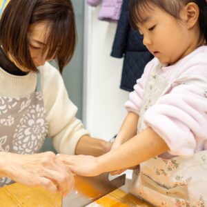 大人に手伝ってもらいながら包丁でうどんを切る女の子。 Girl cutting the udon dough with a knife, assisted by an adult. 手打ちうどん作り体験教室 2026年1月 A6408467-1.jpg – イリコスキー製麺所