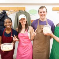 完成したうどんを容器に入れて、エプロン姿で並ぶ記念写真。 / A group photo in aprons holding containers of handmade udon noodles. 手打ちうどん作り体験教室 2026年2月 iricosky-group-photo-handmade-udon-containers.jpg – イリコスキー製麺所