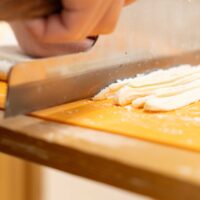 麺切り包丁を当ててうどんを細く切っている手元のアップ。 / A close-up of hands using a udon knife on a cutting board to slice noodles thinly. 手打ちうどん作り体験教室 2026年2月 udon-cutting-closeup-knife-thin-noodles.jpg – イリコスキー製麺所