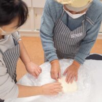  踏みの合間にうどん生地を折りたたんでいる２人の参加者。Two participants folding udon dough between stepping
  rounds.