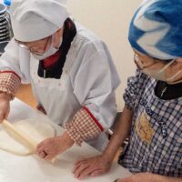 白い割烹着の女性が麺棒でうどん生地を延ばしている。A woman in a white apron rolling out udon dough with a rolling pin. 出張手打ちうどん作り体験 2026年3月20日 A6409915-udon-class-rolling-dough-white-apron-1.jpg – イリコスキー製麺所
