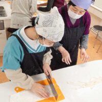 緑のセーターの女の子が包丁でうどんを切っている。A girl in a green sweater cutting udon noodles with a kitchen knife. 出張手打ちうどん作り体験 2026年3月20日 A6409943-udon-class-girl-cutting-noodles-green-vneck-1.jpg – イリコスキー製麺所