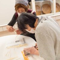 の女性が切ったうどんを確認しながら麺切りしている。A woman in a striped shirt cutting udon noodles on a
  cutting board.