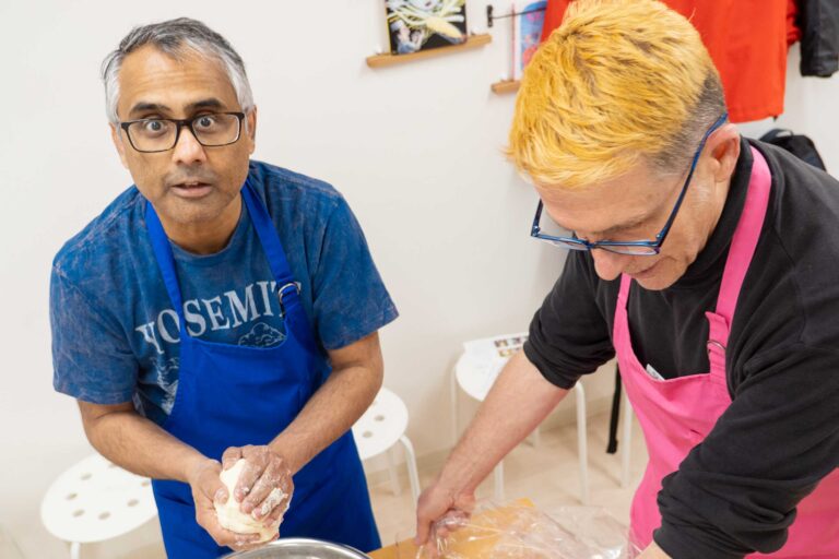 イリコスキーうどん作り体験教室で粉と水を混ぜた生地をひとかたまりにまとめている海外ゲスト2人。青エプロンの方がカメラに目を向けている。 手打ちうどん作り体験教室 2026年1月 udon-class-dough-gathering-two-guests-osaka.jpg – イリコスキー製麺所