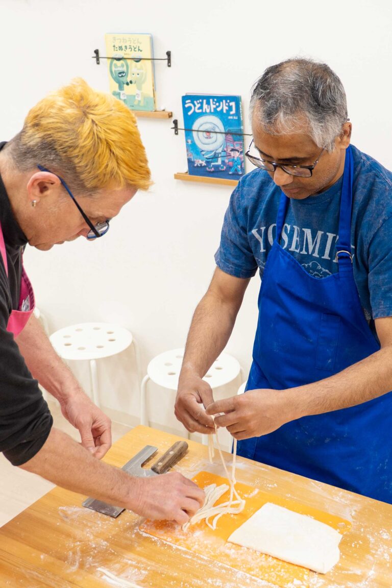 イリコスキーうどん作り体験教室で麺切りを終えた直後、2人の海外ゲストがテーブルの上で麺を引き出している場面。包丁とオレンジのまな板、打ち粉が並ぶ。 手打ちうどん作り体験教室 2026年1月 udon-class-noodle-cutting-two-guests-osaka.jpg – イリコスキー製麺所