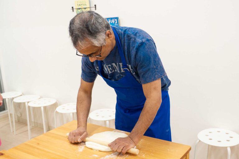 イリコスキーうどん作り体験教室で麺棒を両手で押さえながら生地を延ばす海外ゲスト。打ち粉の舞うテーブルで集中した横顔が印象的。 手打ちうどん作り体験教室 2026年1月 udon-class-rolling-dough-guest-profile-osaka.jpg – イリコスキー製麺所