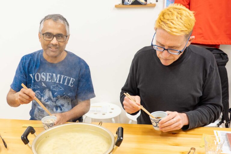 イリコスキーうどん作り体験教室で自作うどんを前に笑顔を見せる海外ゲスト2人。大鍋と薬味の並ぶテーブルを囲み、温かな雰囲気で体験を締めくくっている。 手打ちうどん作り体験教室 2026年1月 udon-class-tasting-smiling-guests-finish.jpg – イリコスキー製麺所