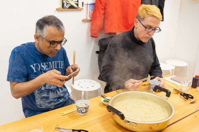 イリコスキーうどん作り体験教室で手打ちうどんを試食する海外ゲスト2人。湯気の立つ鍋を前に、1人はスマートフォンで撮影し、もう1人は箸でうどんをすくっている。 手打ちうどん作り体験教室 2026年1月 udon-class-tasting-steam-pot-two-guests.jpg – イリコスキー製麺所