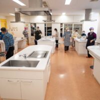 出張うどん体験教室の会場全景。調理実習室に参加者が集まり、各テーブルで作業の準備をしている。Participants
  gathering at the cooking classroom for an outgoing udon-making workshop.