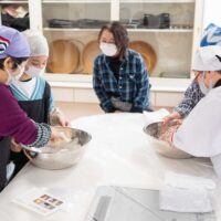 ３人の女性がボウルの中で小麦粉と水を混ぜ合わせている。Participants mixing flour and water in large bowls at the
  start of the udon-making process.