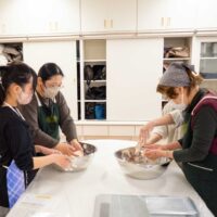 親子と女性がボウルの中でうどん生地を混ぜている。A mother-daughter pair and another participant mixing udon dough
  in bowls.