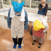 青いバンダナの男性がビニール袋に入れたうどん生地を足で踏んでいる。A man stepping on udon dough in a plastic bag
  to knead it with his feet.