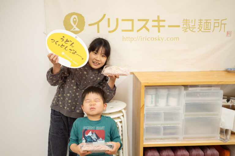 イリコスキー製麺所のバナーの前で、完成したうどんとフォトプロップスを持って記念撮影する子どもたち。