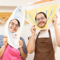 イリコスキー製麺所の顔はめパネルで笑顔の記念撮影。女性はいりこのパネル、男性はうどんのパネルに顔を出してにっこり。 手打ちうどん作り体験教室 2026年5月 udon-class-face-panel-sardine-noodle-bowl-portrait.jpg – イリコスキー製麺所