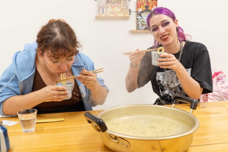 2人が並んで着席し、できたての熱々うどんを箸でつまみながら試食している。棚にはマンガ風の絵本が並んでいる。 手打ちうどん作り体験教室 2026年2月 udon-class-tasting-chopsticks-camera-smile.jpg – イリコスキー製麺所