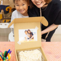 父の日のプレゼントボックスの前で笑顔を見せる女の子とお母さん。箱の蓋には体験中の写真が貼られ、中にはできたてのうどんとつゆが詰められている。 手打ちうどん作り体験教室 2026年6月 udon-class-fathers-day-gift-box-mother-daughter-smile.jpg – イリコスキー製麺所