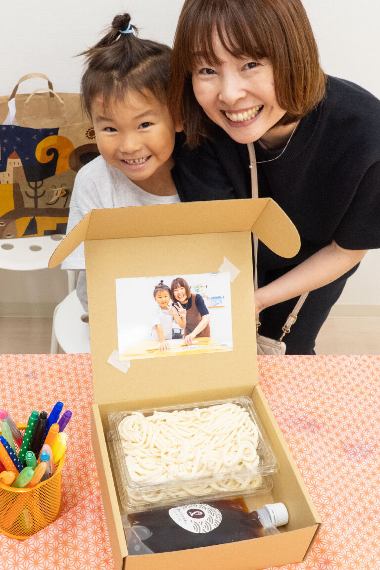 父の日のプレゼントボックスの前で笑顔を見せる女の子とお母さん。箱の蓋には体験中の写真が貼られ、中にはできたてのうどんとつゆが詰められている。 手打ちうどん作り体験教室 2026年3月 udon-class-fathers-day-gift-box-mother-daughter-smile.jpg – イリコスキー製麺所