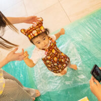 帽子とエプロン姿の赤ちゃんが、手を支えてもらいながらうどん生地を足で踏んでいる。A baby in a hat and apron stepping on udon dough while holding an adult's hands. 出張手打ちうどん作り体験 2026年8月8日 20260808-2.jpg – イリコスキー製麺所